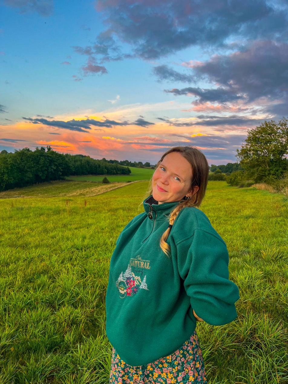 Hanne staat glimlachend in een weids groen veld bij zonsondergang met felgekleurde wolken
